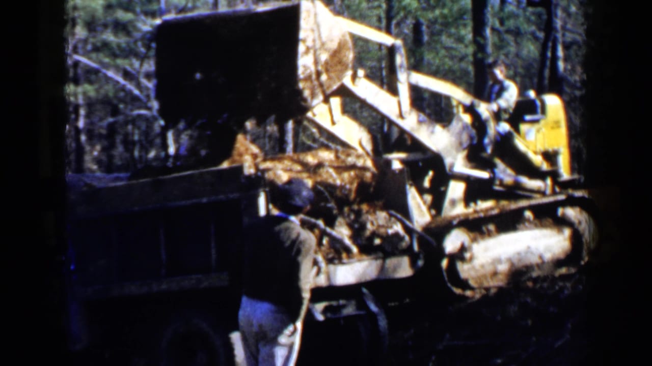 Yellow loader dumping dirt into a truck bed while a man stands nearby in woods.