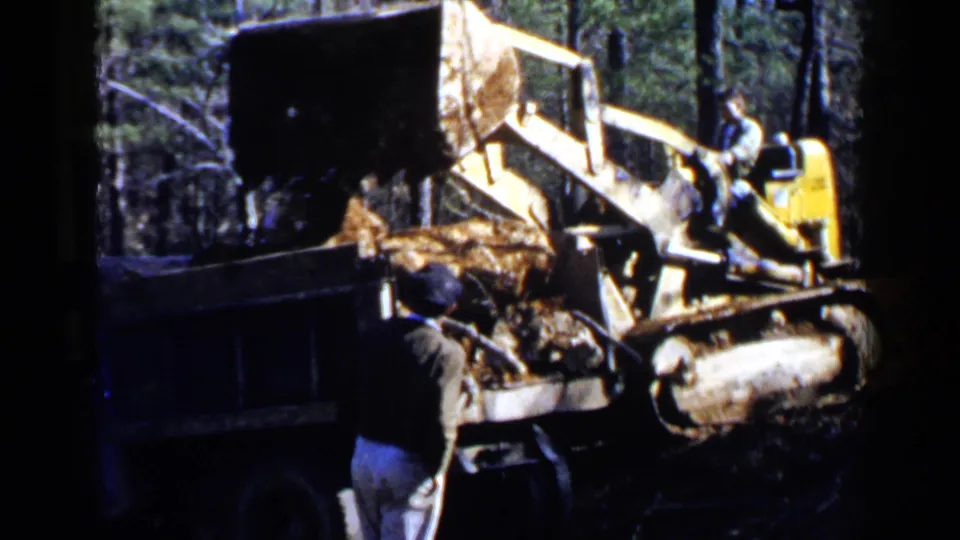 1957 Birmingham-area 8mm frame showing a yellow tracked loader tipping dirt into a dump truck as a man observes in wooded terrain.