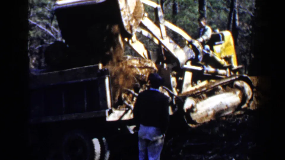 Yellow construction loader empties soil into a dump truck in wooded Birmingham, Alabama footage from 1957, with one adult figure nearby.