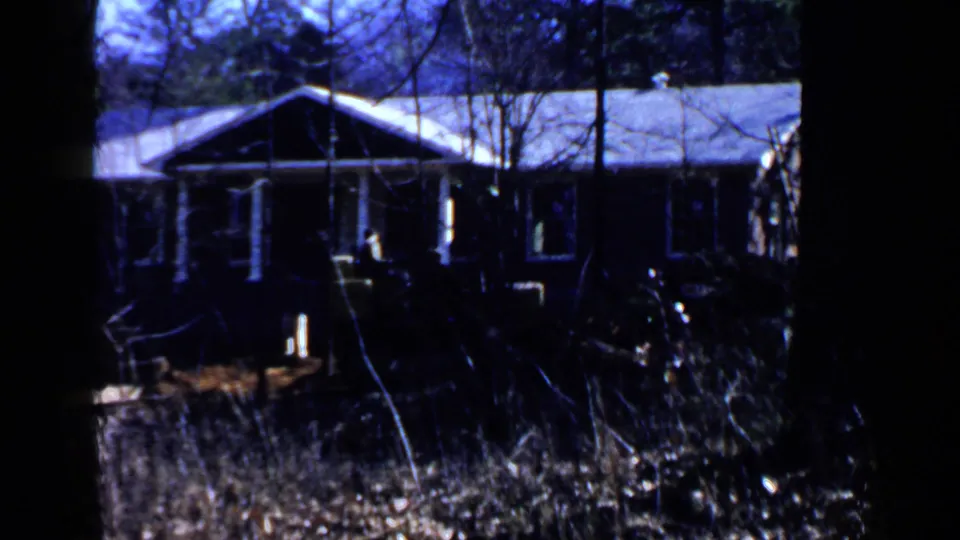 1957 Birmingham-area home-movie frame showing a small cabin-like building through trees after the construction machinery shots.