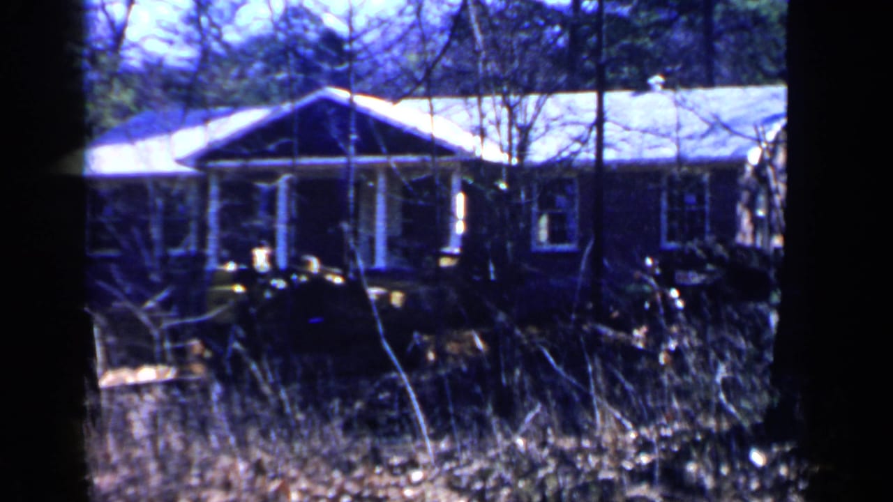 Small single-story house behind tall grass and trees, with a dark vehicle or equipment in front.