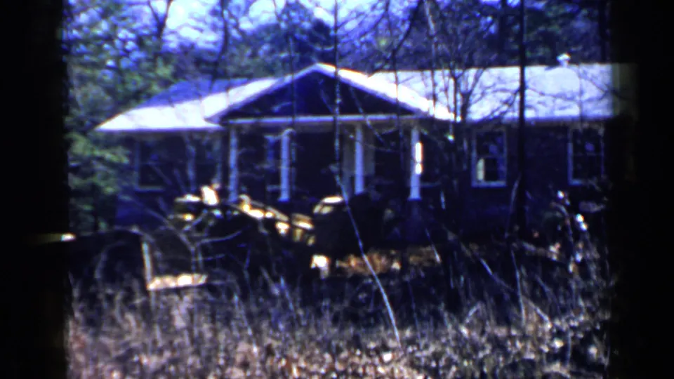 1957 Birmingham-area home-movie frame of a small house with a pale roof, porch posts, surrounding woods, and equipment or vehicle shapes in front.