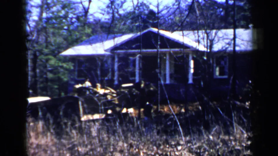 1957 Birmingham-area 8mm frame with the porch centered, bare branches crossing the view, tall grass below, and work equipment or a vehicle at left.