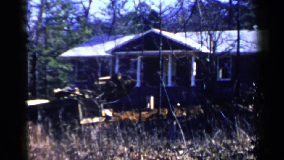 1957 Birmingham-area frame showing the dark house front, white porch supports, bare trees, tall grass, and partial vehicle or work equipment at the edge.