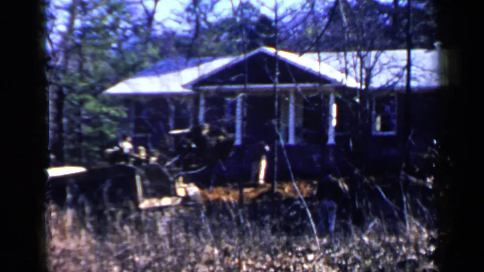 1957 Birmingham home-movie frame with a person operating a yellow bulldozer near a brown house, partially hidden by foreground brush.