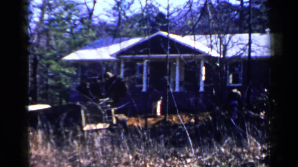 Final 1957 Birmingham frame showing the yellow bulldozer near a brown residential exterior, with trees, brush, and tall grass across the view.