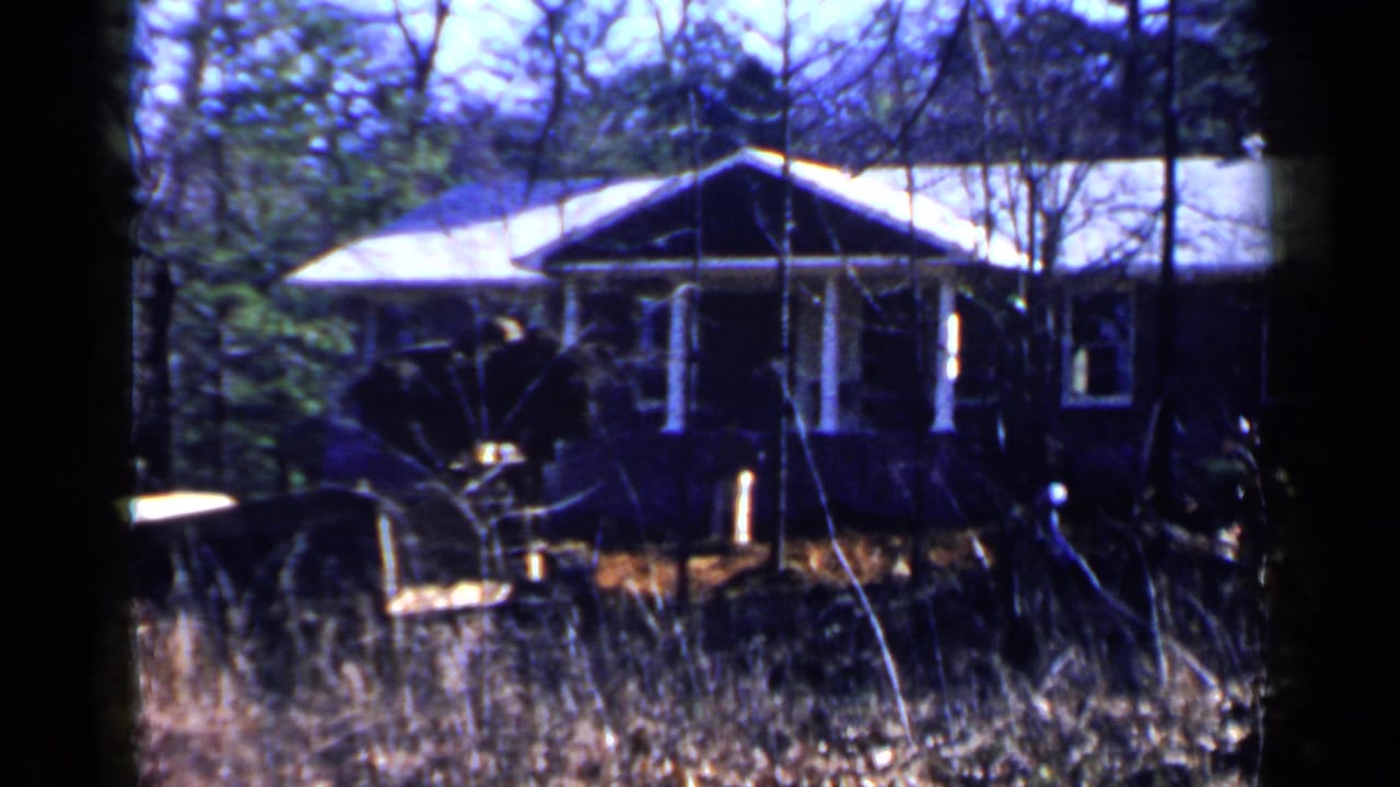 Yellow loader near a porch-front house in tall grass and trees.