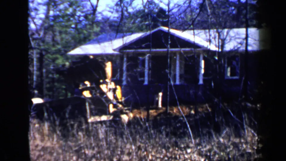 1957 Birmingham home-movie still showing the yellow loader working near overgrown vegetation by the house.