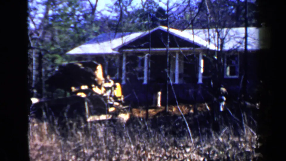1957 Birmingham frame showing a compact loader near a modest house, viewed through a wooded yard.
