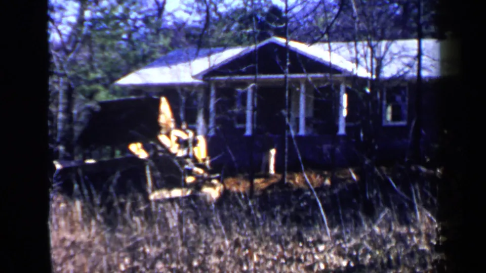1957 Birmingham frame with a yellow construction machine foregrounded against a small house and leafless trees.