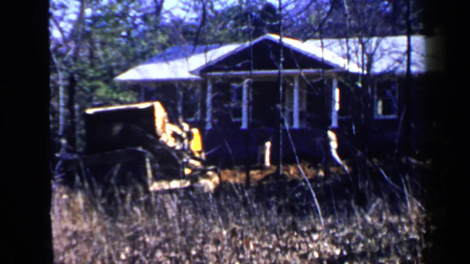Home-movie frame from Birmingham in 1957 showing a yellow construction vehicle beside a wooded residential house.