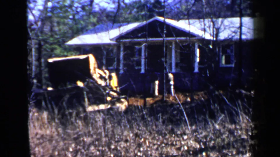 1957 Birmingham 8mm frame of a yellow construction vehicle near a single-story house in a wooded lot.