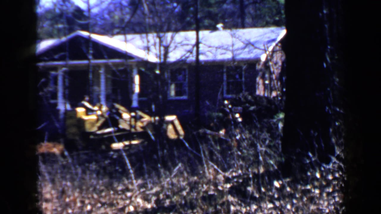 Yellow front-end loader in brush near a ranch-style house.