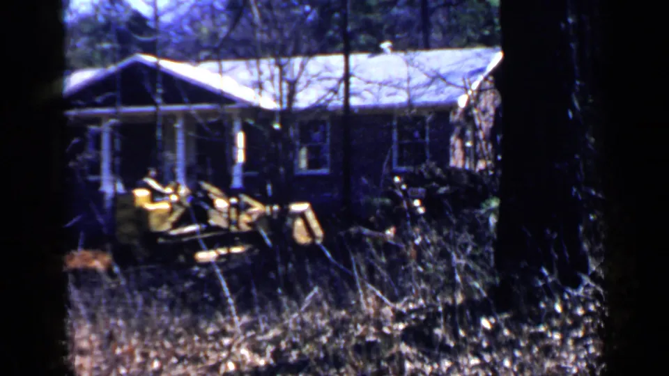1957 Birmingham-area home-movie frame showing a yellow loader working near a low house in a wooded lot.