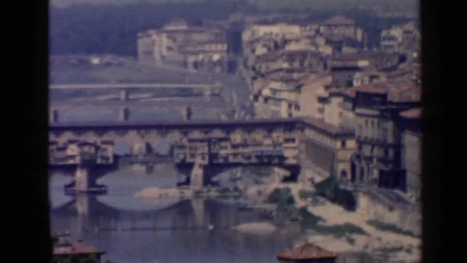 1960 8mm view of Ponte Vecchio over the Arno River, with Florence buildings, bridge arches, and distant hillside visible.