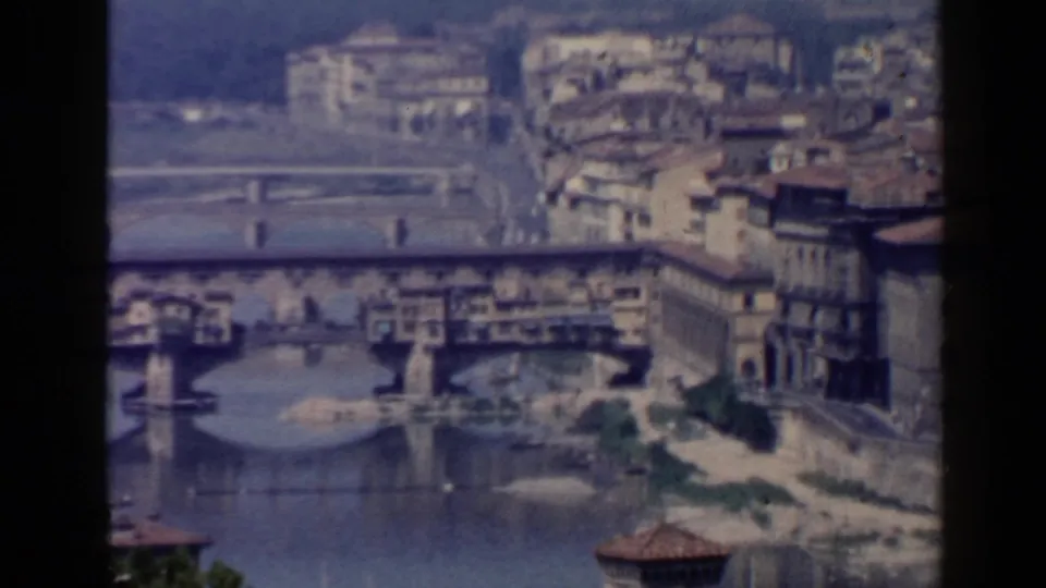 1960 8mm frame showing Ponte Vecchio across the Arno, with shop-lined bridge structure and dense urban buildings nearby.