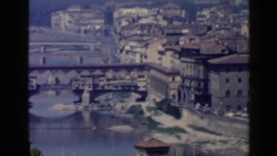1960 8mm view of Ponte Vecchio over the Arno, with a prominent riverside building, bridge arches, and tiled roofs visible.