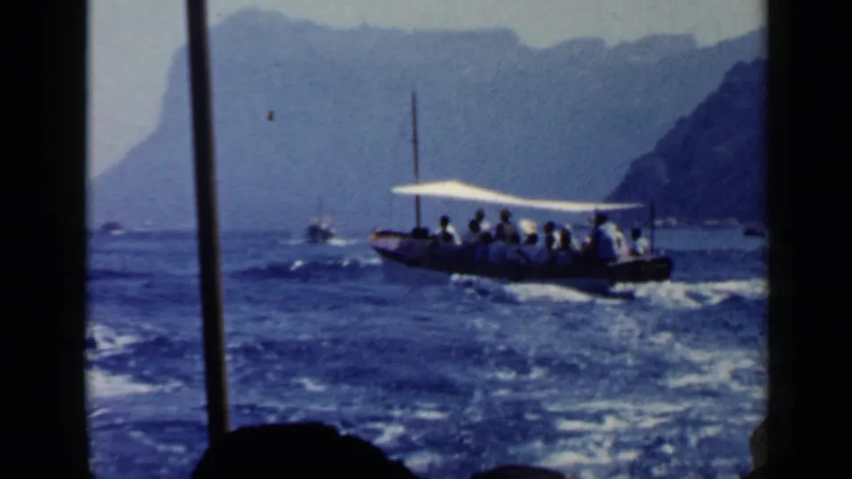 1960 Italy travel frame showing a canopy tour boat crossing blue water near a hazy mountainous shoreline.