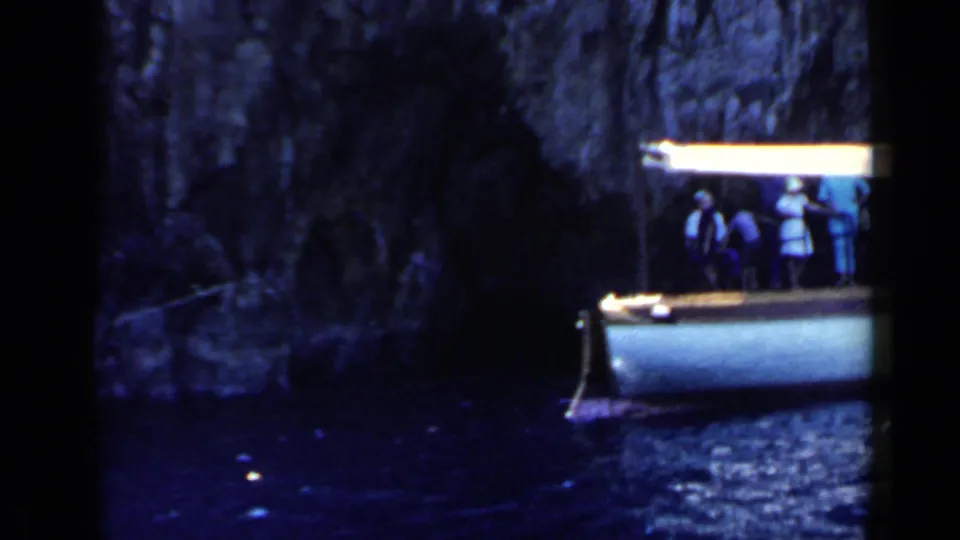 Small leisure boat with people aboard near a shadowed rocky cove wall in 1960 home-movie footage from the Rome, Italy metadata location.