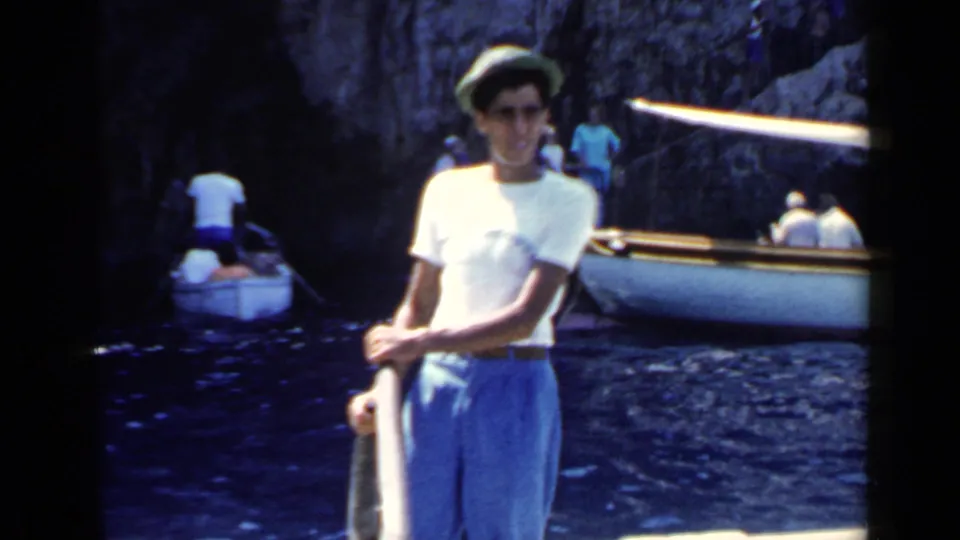 Young man in a light shirt, blue pants, and green hat stands on a boat while other tourists sit in small boats in a rocky cove.