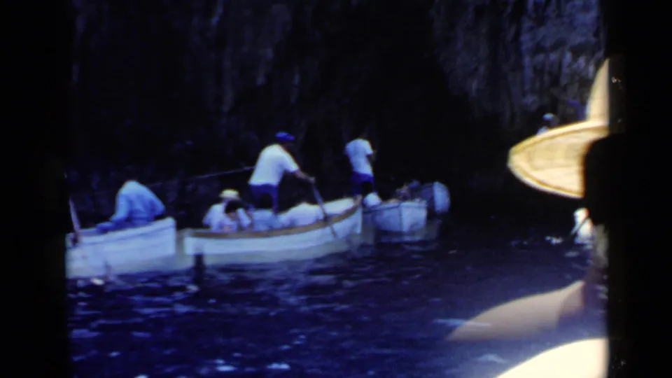 Several small boats with seated people move through dark blue water beside a shadowed rocky wall, with part of a hat visible at the frame edge.