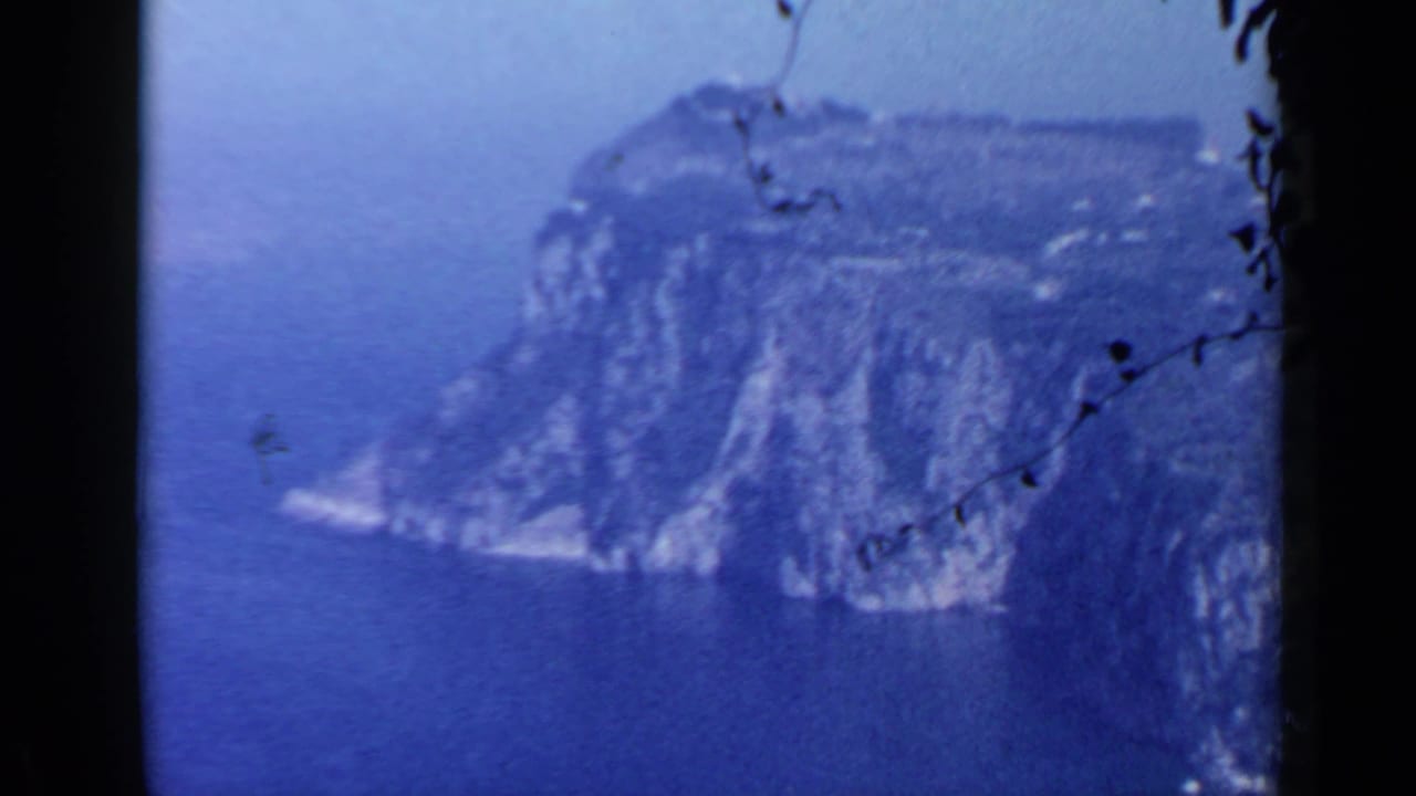 1960 8mm view near Rome showing a steep coastal cliff above deep blue sea, framed by dark film edges and branches.