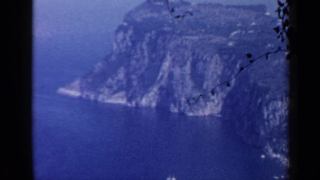 1960 coastal view near Rome with a rocky headland, blue sea, and visible 8mm frame edges.