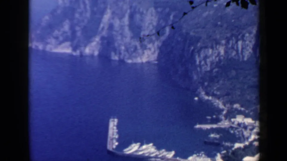 1960 8mm harbor view near Rome, showing a curved pier and boats below steep coastal cliffs.