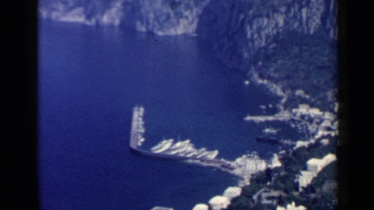 Elevated view of a curved harbor pier, docked boats, blue water, and cliffside buildings.