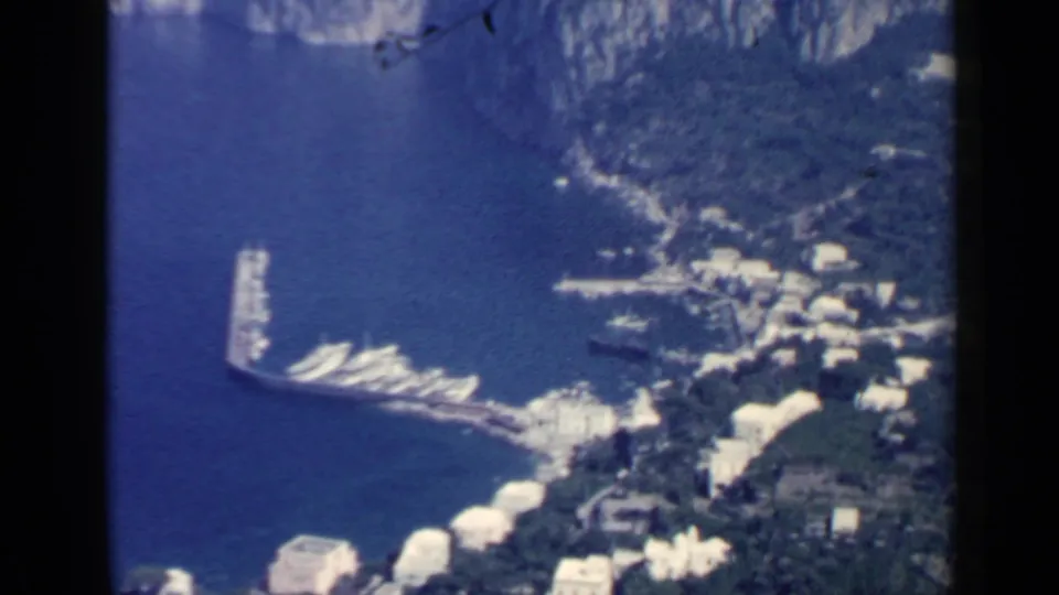 Elevated 1960 Italy coastal view showing a harbor pier, small boats, hillside buildings, and rocky shoreline.