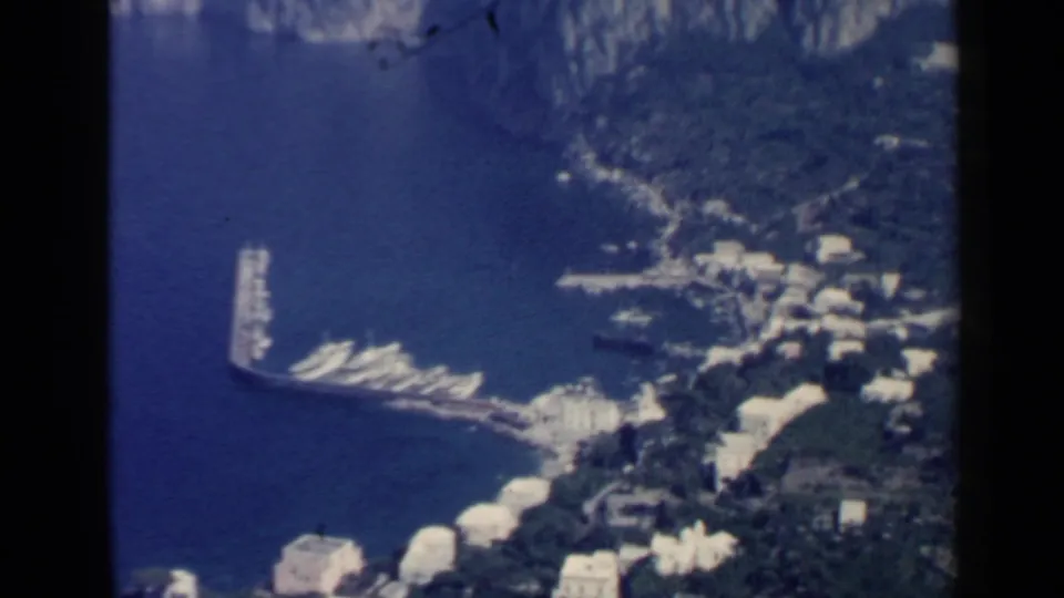 Soft 8mm frame of a coastal Italian harbor, with boats along a curved pier and white buildings near the water.