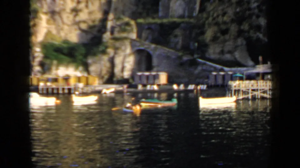 1960 Italian harbor scene with moored boats, a cliffside arched tunnel, quay structures, and a waterside platform.