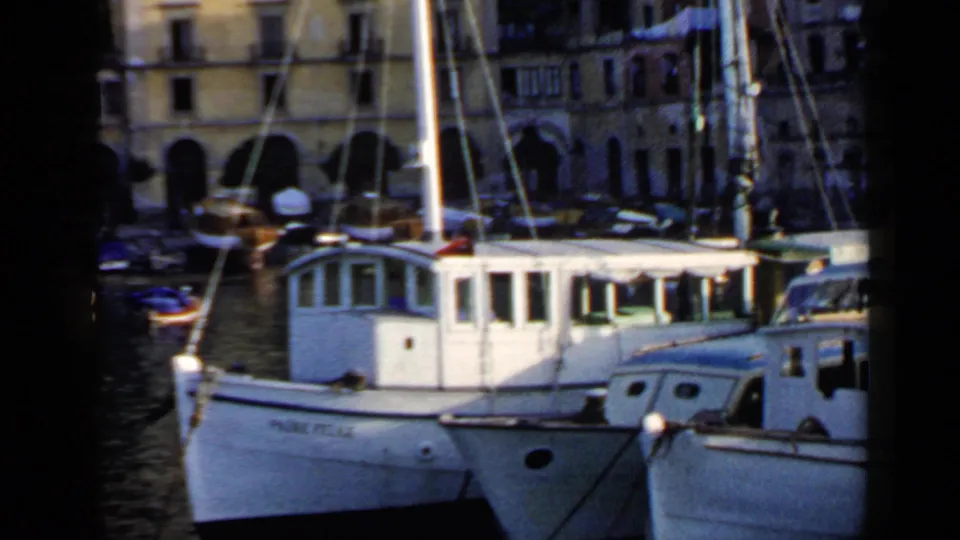 1960 Italian harbor view with the Fabio Felice and another white boat moored before stone buildings with arched openings.