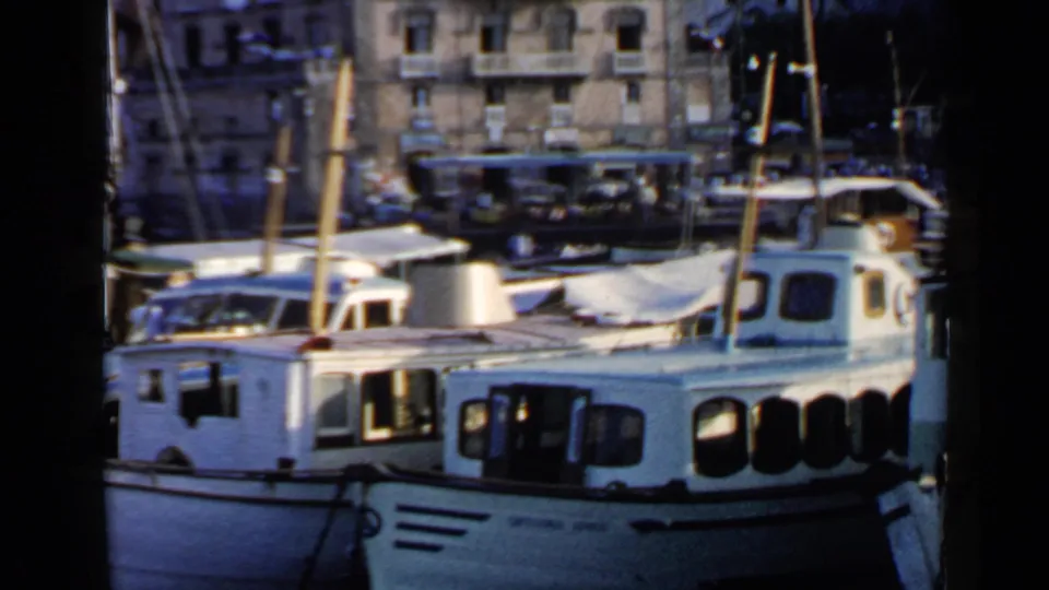 1960 8mm view of a Rome, Italy harbor area with white boats moored near waterfront buildings and a bus beyond them.