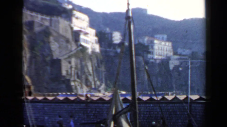 A soft 8mm view of the Rome coastal waterfront shows hillside buildings, a seawall, rigging, and small pedestrians near the shore.