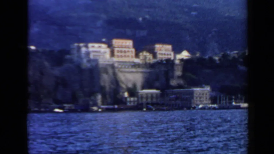1960 8mm Rome travel view of pale waterfront buildings set into a steep hillside above harbor water.