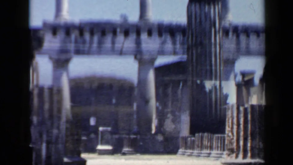 1960s 8mm travel view of Italian ruins with columns, walls, railings, and bright sky, framed by dark film edges.