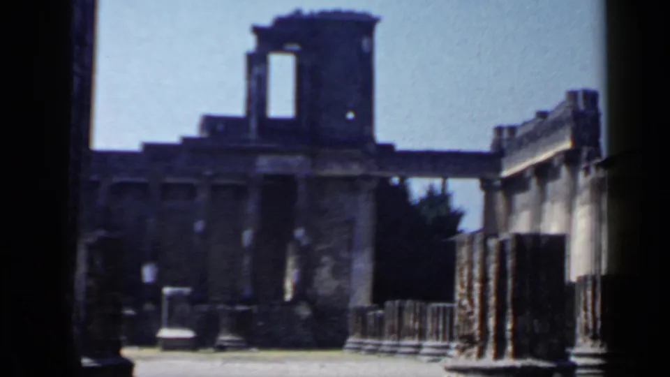 1960s 8mm view of an Italian archaeological ruin with standing columns, broken masonry, railings, and open blue sky.
