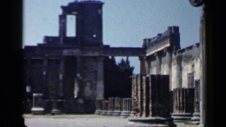 1960s 8mm travel frame showing Italian ruins with a tall column in front, worn masonry, railings, and a shadowed wall opening.