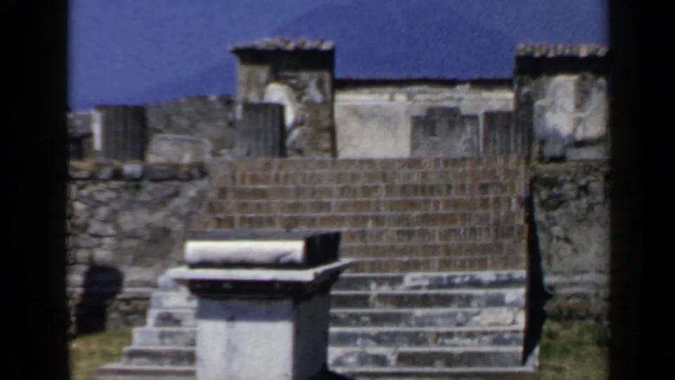 1960 Rome home-movie frame with a closer composition of ancient masonry, staircases, stone block, grass, and blue sky.