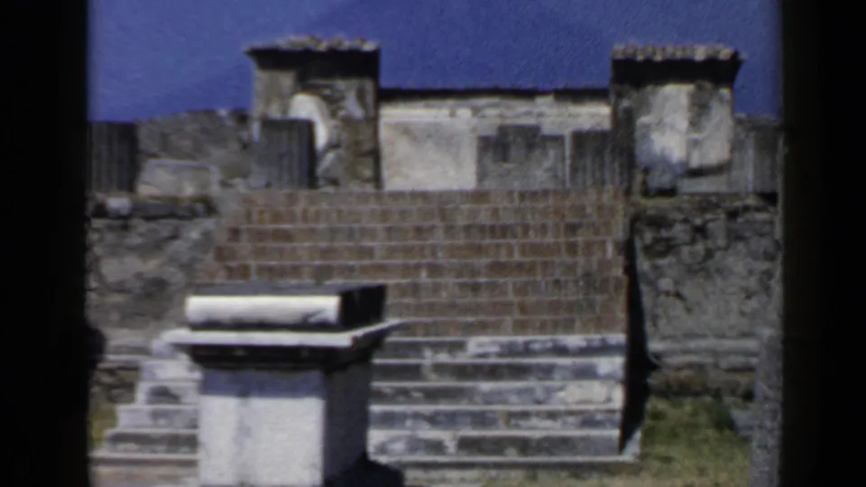 Rome, Italy 1960 8mm frame showing ancient stone steps, a column fragment, masonry walls, grass, and a central rectangular block.