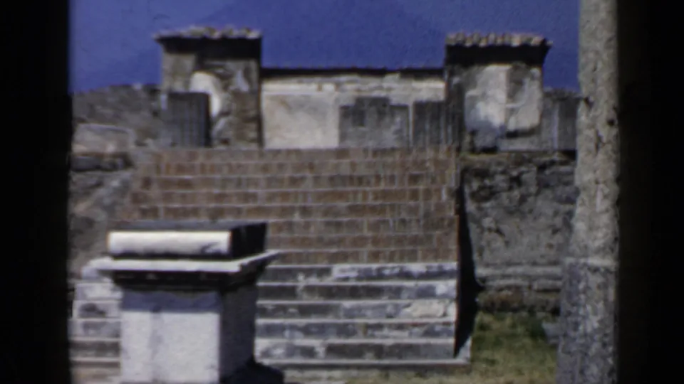 1960 8mm view of sunlit archaeological stone steps, rough masonry walls, and roofline remnants in Italy.