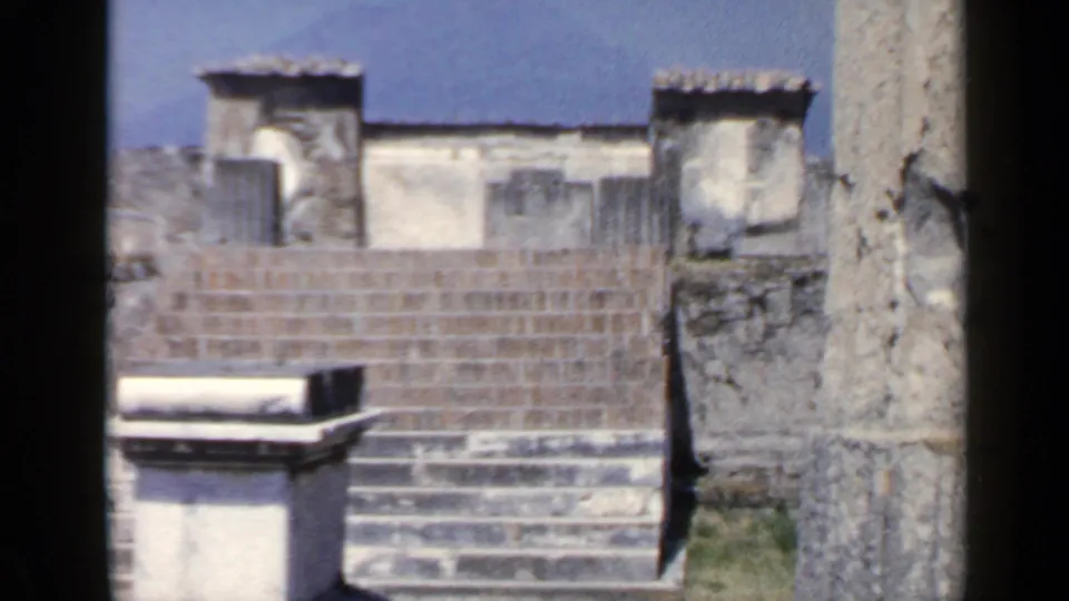 1960 8mm frame showing a sunlit ruin wall, stone staircase, and rectangular block with dark film-edge framing.