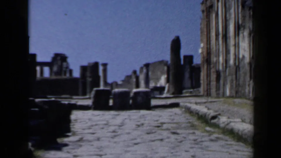 1960 8mm wide view down a stone-paved archaeological street with columns, low blocks, and weathered walls.