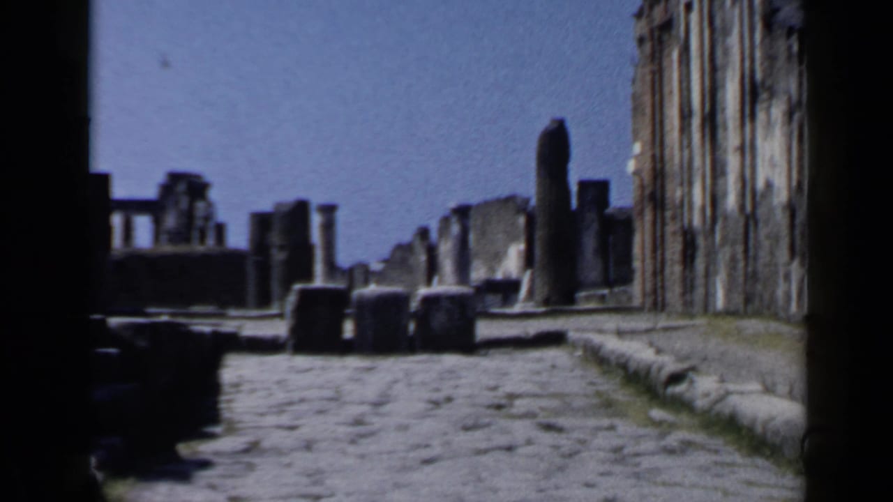 1960 Rome home-movie frame showing a cobblestone path through ancient Roman ruins with columns and walls in sunlight.