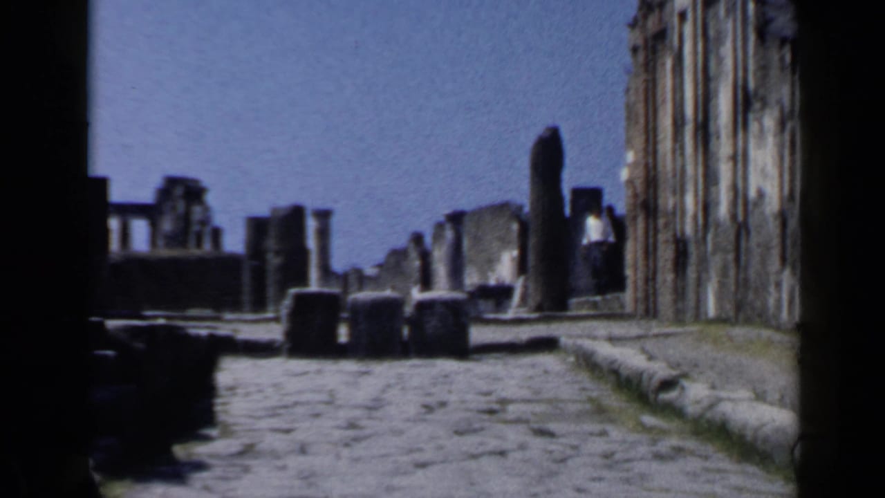 Ancient ruins in Rome appear along a cobblestone street, framed by dark foreground shadows and bright sky.