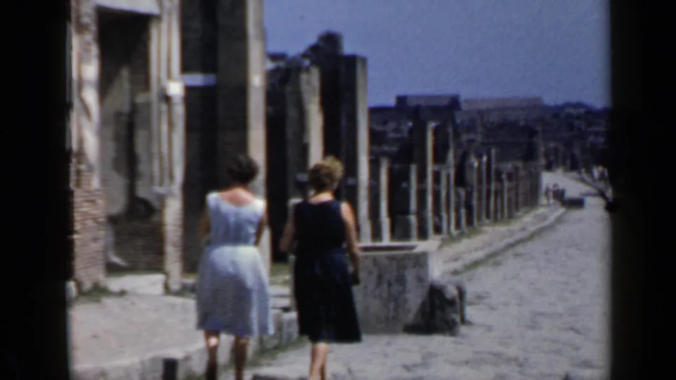 Two women in mid-century dresses walk away through Roman ruins on a long cobblestone street in 1960 Rome.