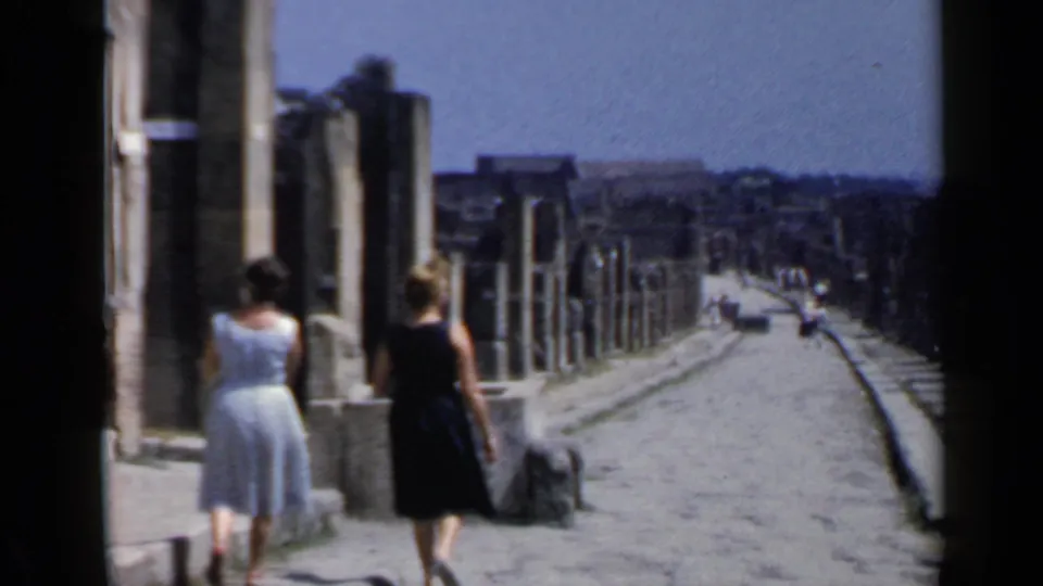 Visitor-shot 8mm frame of two women walking away beside ancient ruins and columns in Rome, Italy.