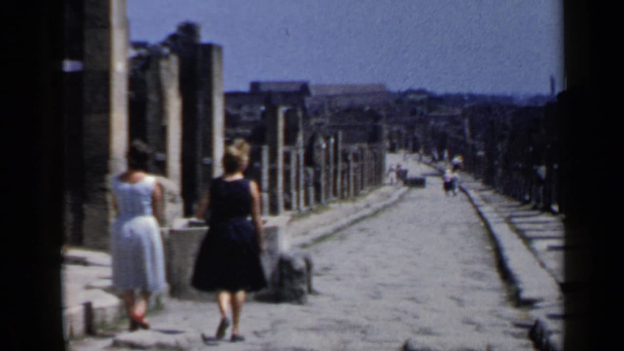 Two women walk along a sunlit stone street lined with columns and old walls.