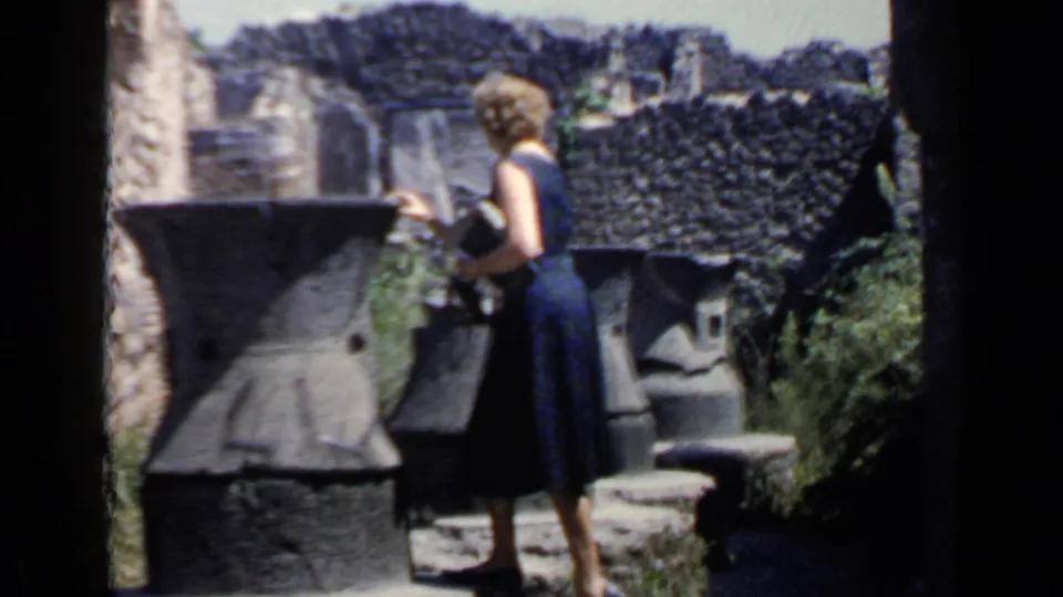 A woman in a dark blue dress examines large stone artifacts at Roman ruins, holding a small item or notebook.